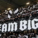 Een volle tribune met zwart-witte supporters die vlaggen omhooghouden.