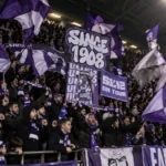 Een groep paars-witte voetbalsupporters die met vlaggen zwaait op de tribune.
