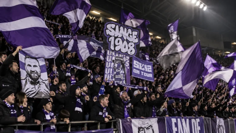 Een groep paars-witte voetbalsupporters die met vlaggen zwaait op de tribune.