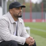 Vincent Kompany zit rustig naast een trainingsveld in een lichte jas en zwarte pet.