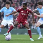 Een Liverpool-speler in donkerrood strijdt om de bal met twee tegenstanders in lichtblauw.