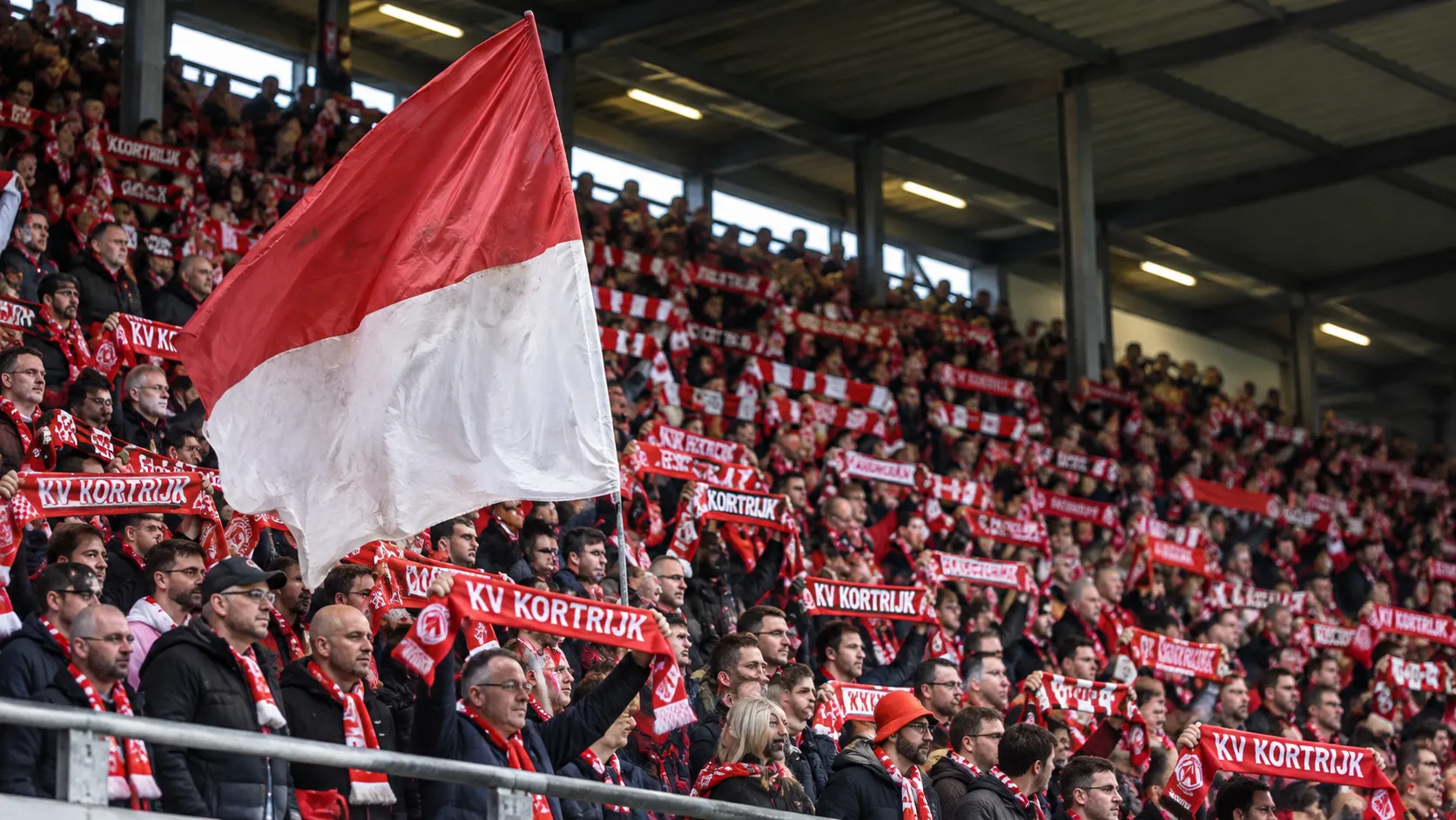 Een groep KV Kortrijk-supporters met rood-witte sjaals op een overdekte tribune.