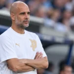 Pep Guardiola in een witte trui langs de rand van een voetbalstadion.