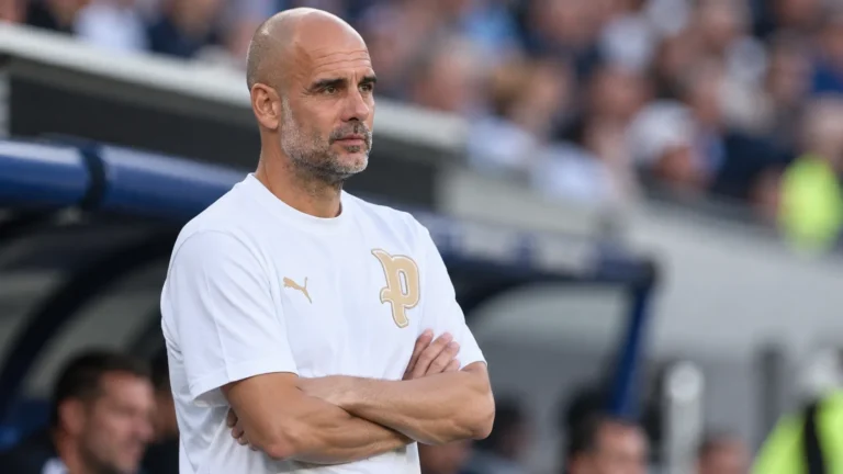 Pep Guardiola in een witte trui langs de rand van een voetbalstadion.