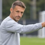 Een niet-spelende clubvertegenwoordiger in grijze trui gebaart langs een trainingsveld.