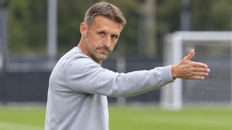 Een niet-spelende clubvertegenwoordiger in grijze trui gebaart langs een trainingsveld.