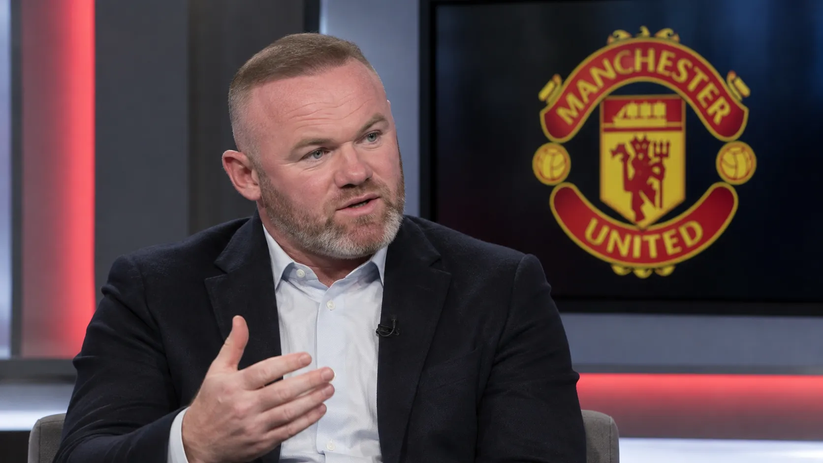 Wayne Rooney spreekt in een studiogesprek, met een Manchester United-embleem op een scherm achter hem.