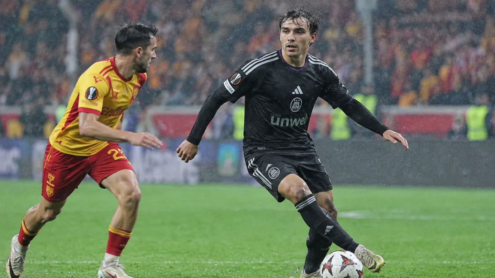Twee voetballers tijdens een regenachtige wedstrijd in een Europees stadion.