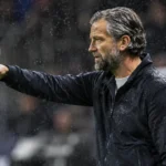 Een coach in donkere jas kijkt bedachtzaam vanaf de zijlijn tijdens een regenachtige voetbalavond.