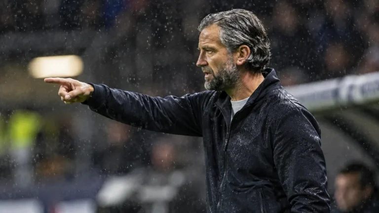 Een coach in donkere jas kijkt bedachtzaam vanaf de zijlijn tijdens een regenachtige voetbalavond.