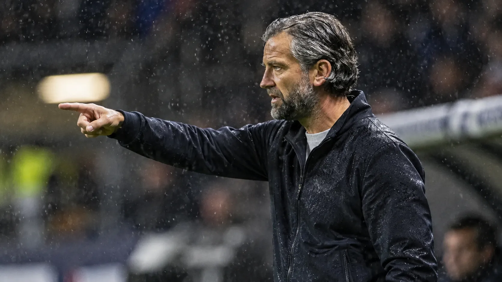 Een coach in donkere jas kijkt bedachtzaam vanaf de zijlijn tijdens een regenachtige voetbalavond.