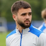 Nicolas Raskin in een wit-blauw trainingsvest op een grasveld.