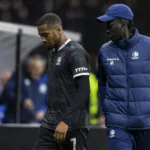 Een KAA Gent-speler in donker tenue loopt langs de zijlijn naast een staflid.