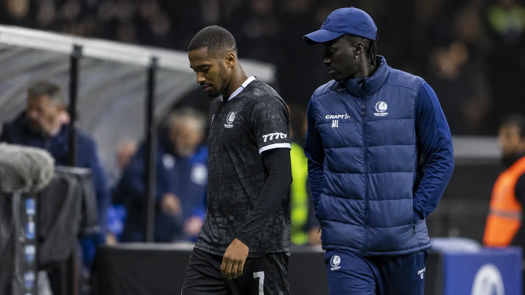 Een KAA Gent-speler in donker tenue loopt langs de zijlijn naast een staflid.