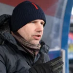 Een clubofficial in donkere winterkleding tijdens een interview naast een overdekte dug-out.