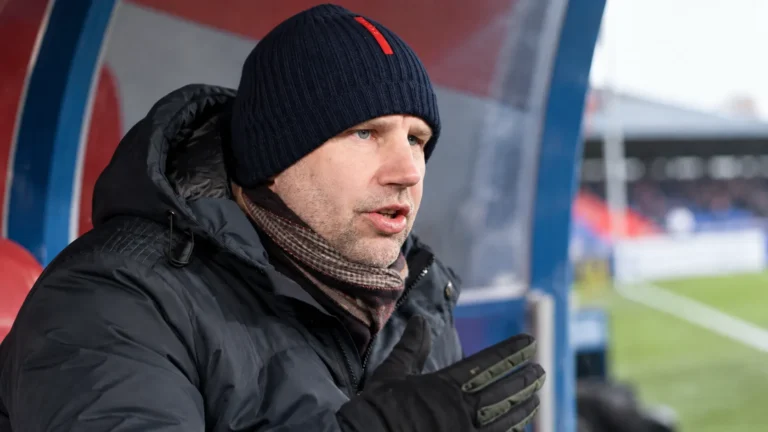 Een clubofficial in donkere winterkleding tijdens een interview naast een overdekte dug-out.