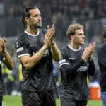 Voetballers en een staflid applaudisseren langs de zijlijn in de regen.