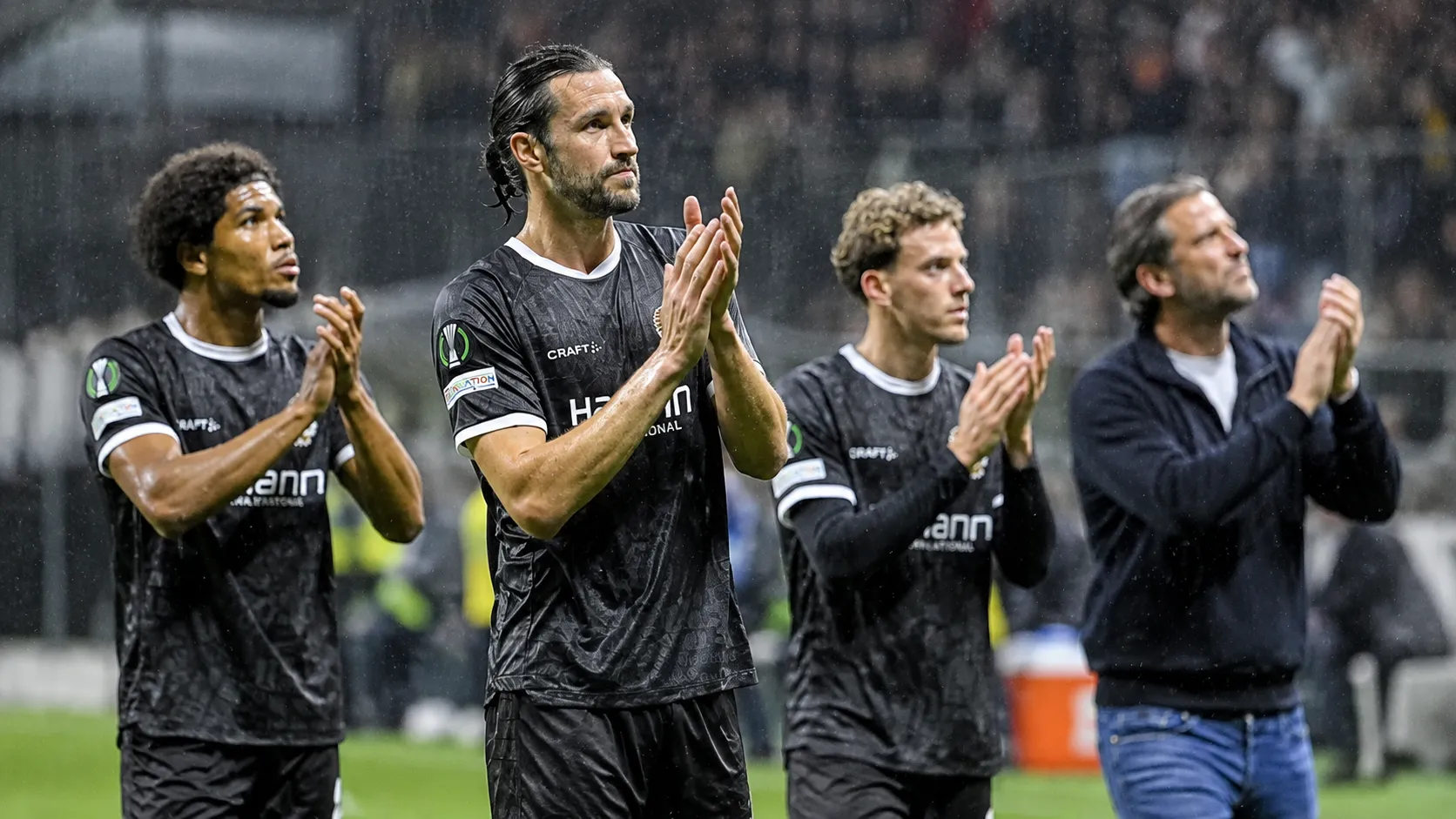 Voetballers en een staflid applaudisseren langs de zijlijn in de regen.