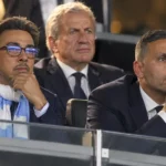 Niet-spelende clubvertegenwoordigers in donkere pakken op de tribune van een voetbalstadion.