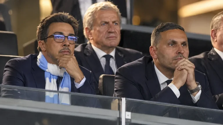 Niet-spelende clubvertegenwoordigers in donkere pakken op de tribune van een voetbalstadion.