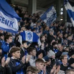 Een collectieve groep Jong Genk-supporters met blauw-witte vlaggen in de tribune.