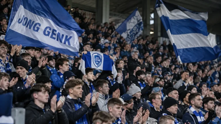 Een collectieve groep Jong Genk-supporters met blauw-witte vlaggen in de tribune.
