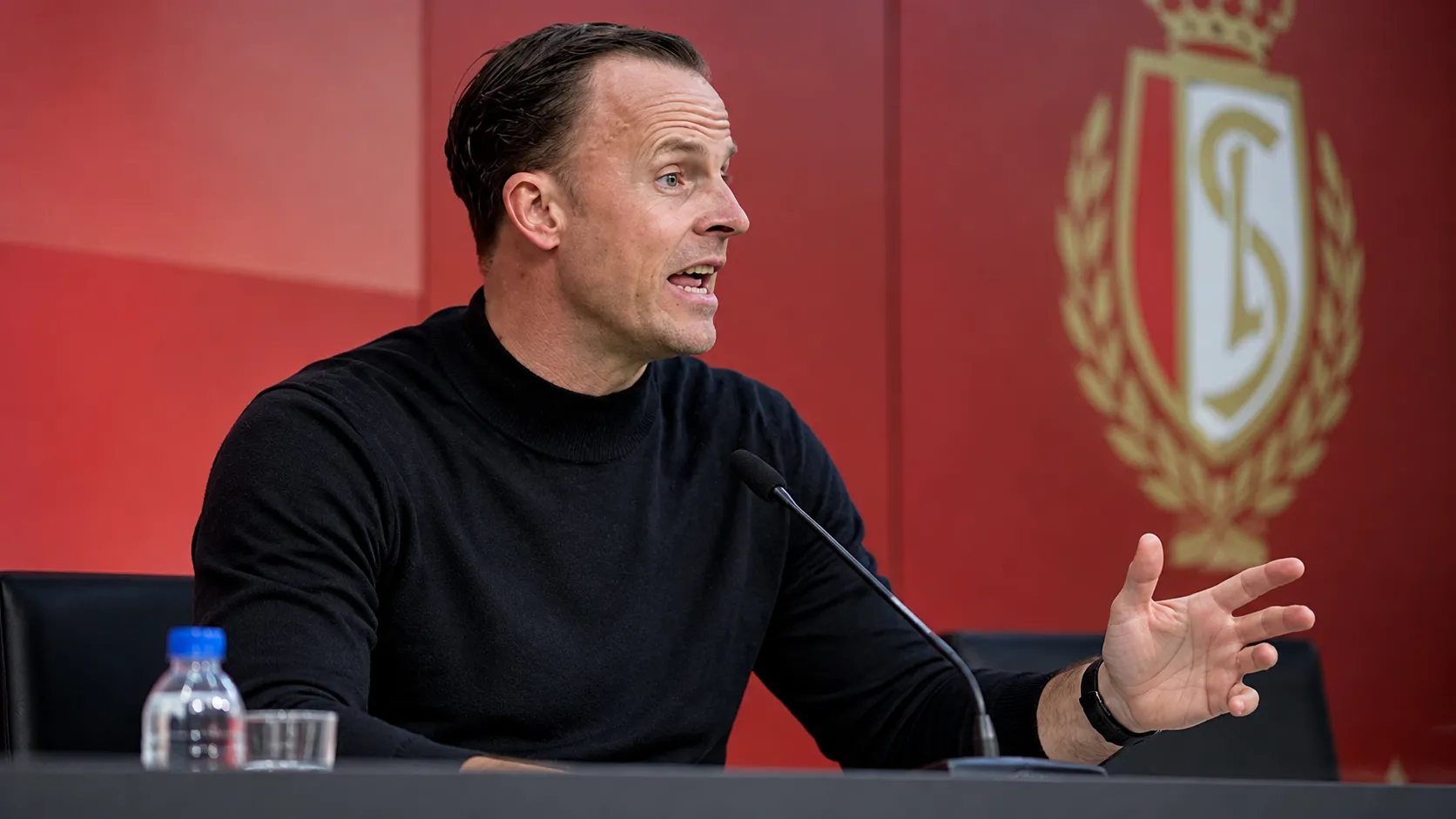 Een Standard-coach in zwarte trui tijdens een persbabbel.