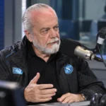 Aurelio De Laurentiis spreekt in een radiostudio met een microfoon, in een donkere Napoli-jas.