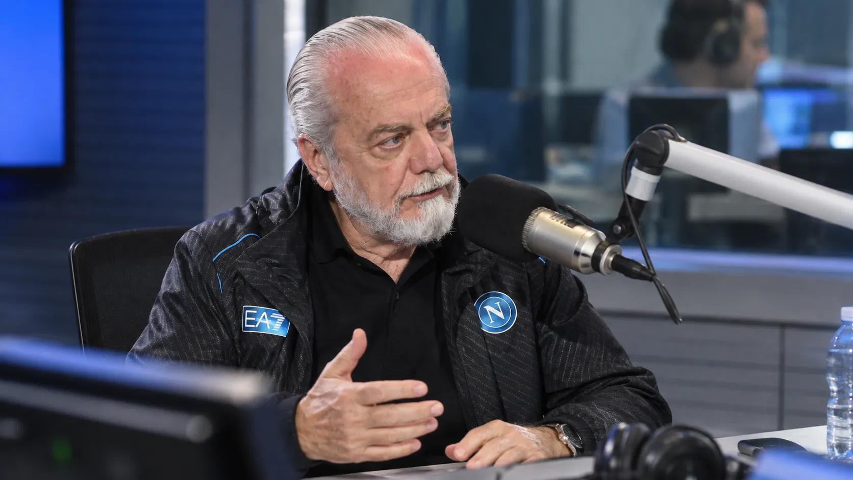 Aurelio De Laurentiis spreekt in een radiostudio met een microfoon, in een donkere Napoli-jas.