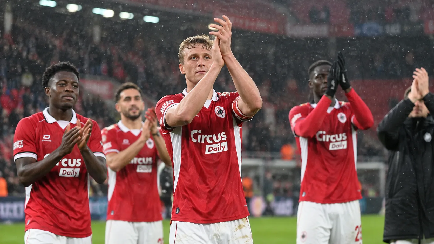 Spelers in rode wedstrijdtenues applaudisseren na afloop op het veld in de regen.