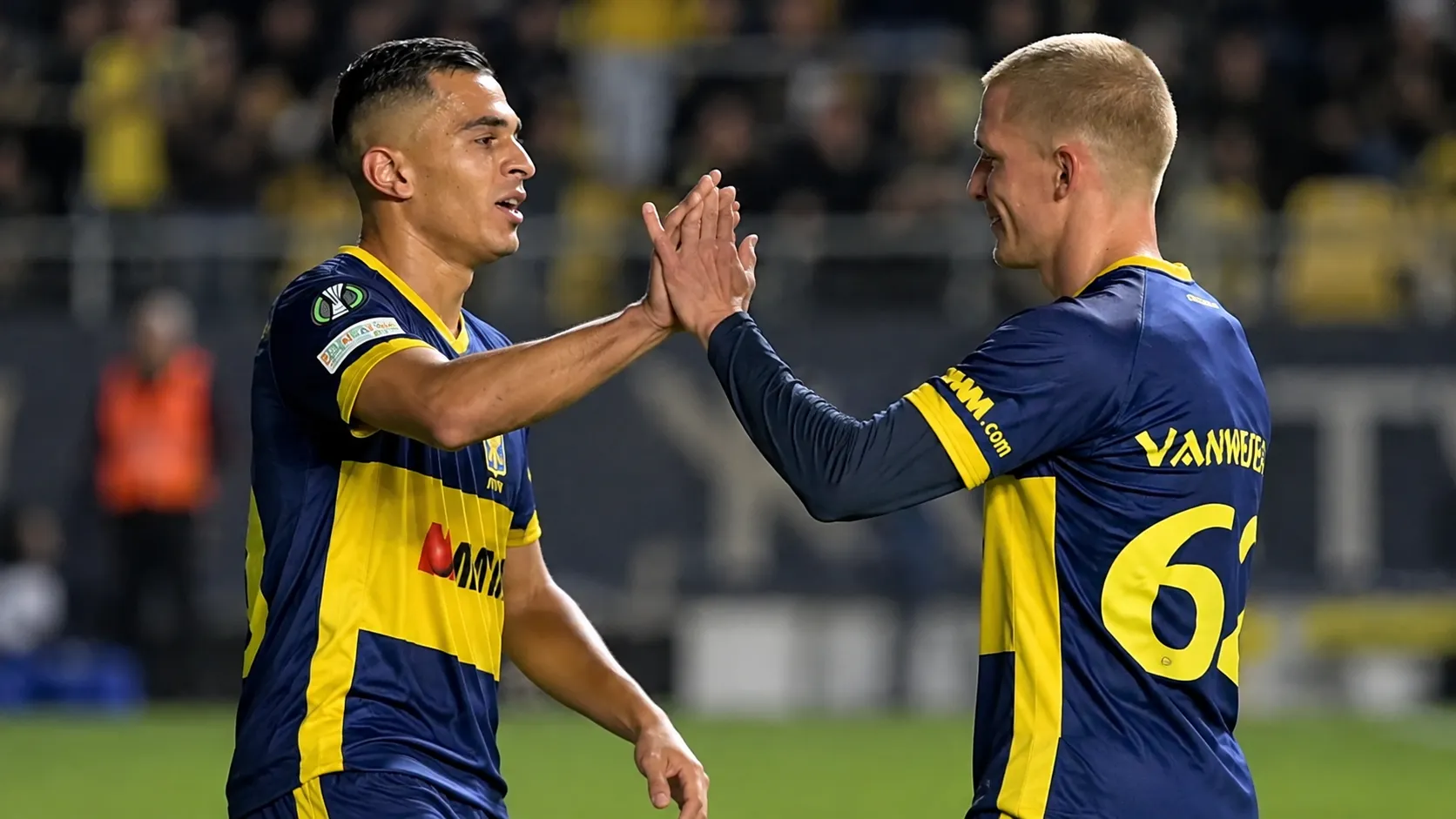 Twee STVV-spelers in blauw-geel tenue moedigen elkaar aan op het veld.