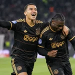 Twee Ajax-spelers in zwart-gouden wedstrijdtenue tijdens een nieuw actiemoment op het veld.