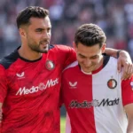 Twee voetballers in rood en rood-wit tenue lopen na een wedstrijd met een arm om elkaar heen.
