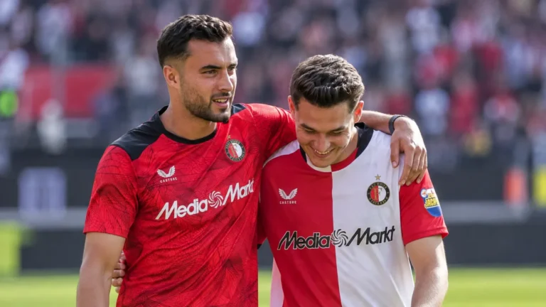 Twee voetballers in rood en rood-wit tenue lopen na een wedstrijd met een arm om elkaar heen.