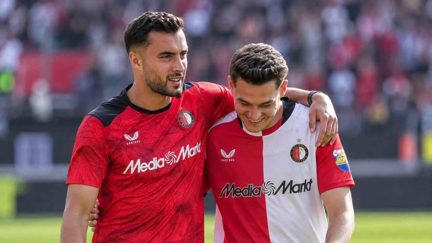 Twee voetballers in rood en rood-wit tenue lopen na een wedstrijd met een arm om elkaar heen.