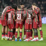 Een groep voetballers in rood-groen gestreepte tenues vormt een huddle op het veld.
