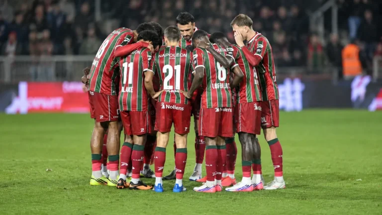 Een groep voetballers in rood-groen gestreepte tenues vormt een huddle op het veld.