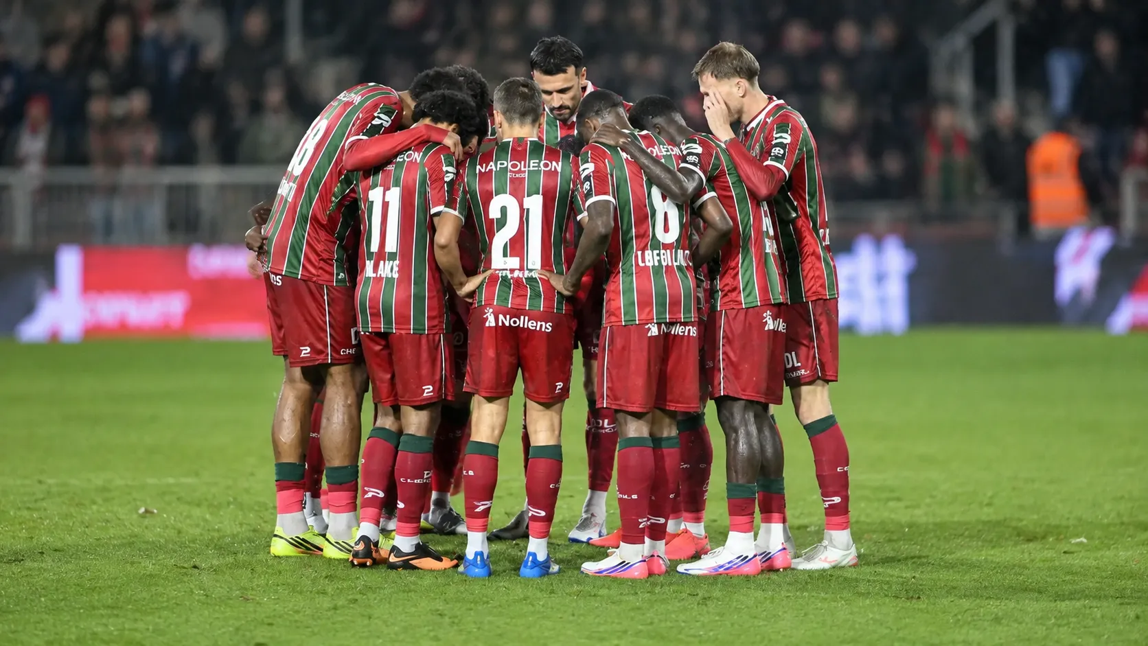Een groep voetballers in rood-groen gestreepte tenues vormt een huddle op het veld.
