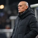 Jürgen Streppel in een donkere jas langs de zijlijn van een voetbalveld.