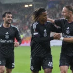 Drie voetballers in donkere tenues vieren samen op een regenachtig voetbalveld.