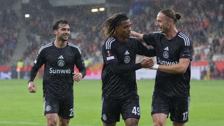 Drie voetballers in donkere tenues vieren samen op een regenachtig voetbalveld.