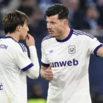 Twee voetballers in witte shirts met paarse details overleggen op het veld.