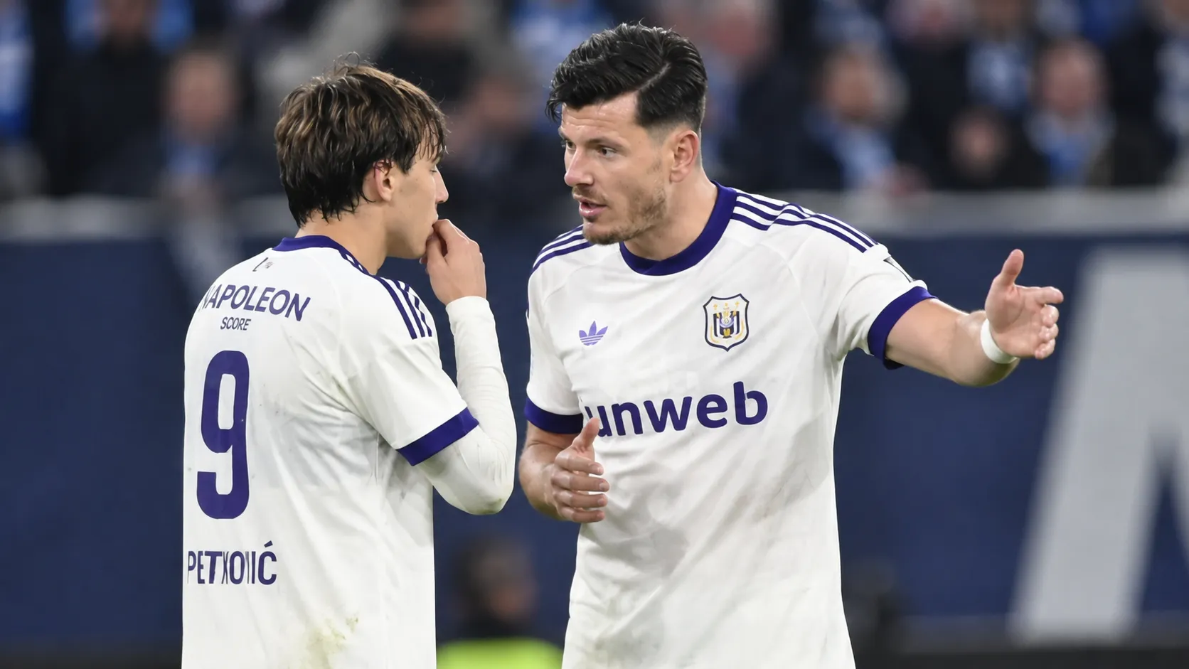 Twee voetballers in witte shirts met paarse details overleggen op het veld.