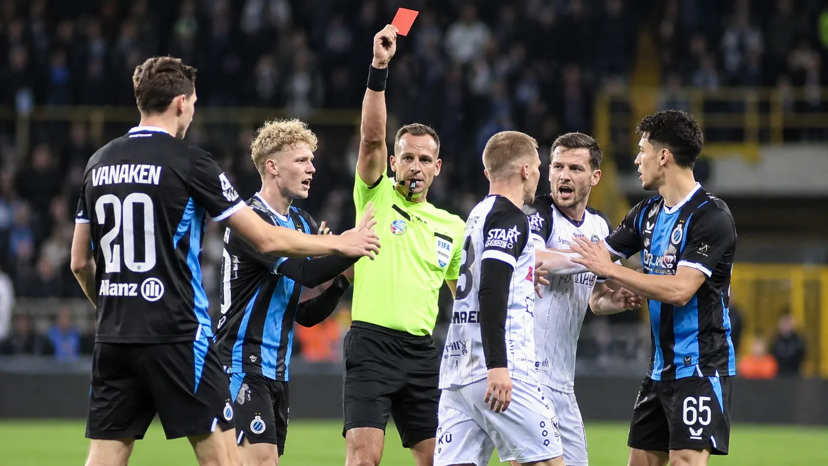 Scheidsrechter toont een rode kaart tussen spelers in zwart-blauwe en witte wedstrijdtenues.