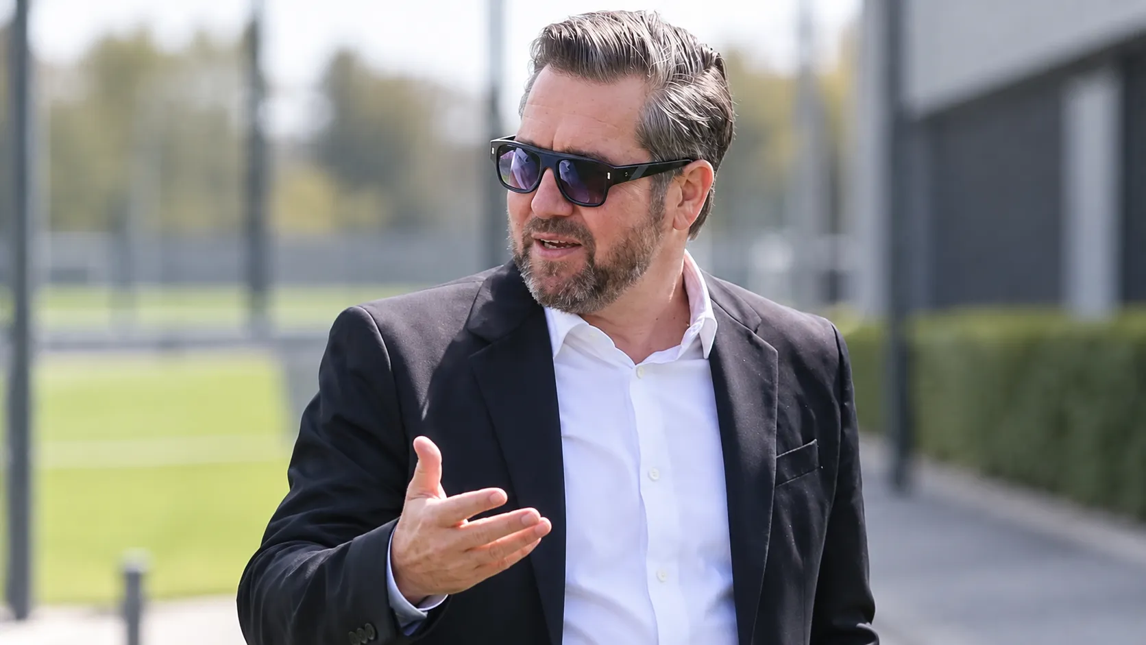 Een niet-spelende voetbalofficial in pak staat buiten in een professionele voetbalomgeving.