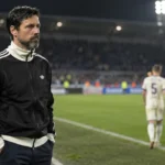 Een coach in zwarte sportjas staat na de wedstrijd langs de zijlijn van een verlicht stadion.