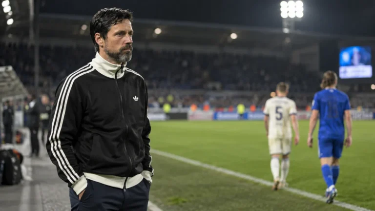 Een coach in zwarte sportjas staat na de wedstrijd langs de zijlijn van een verlicht stadion.