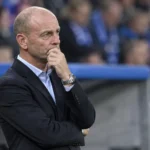 Een KRC Genk-coach in donker pak staat geconcentreerd langs de zijlijn.
