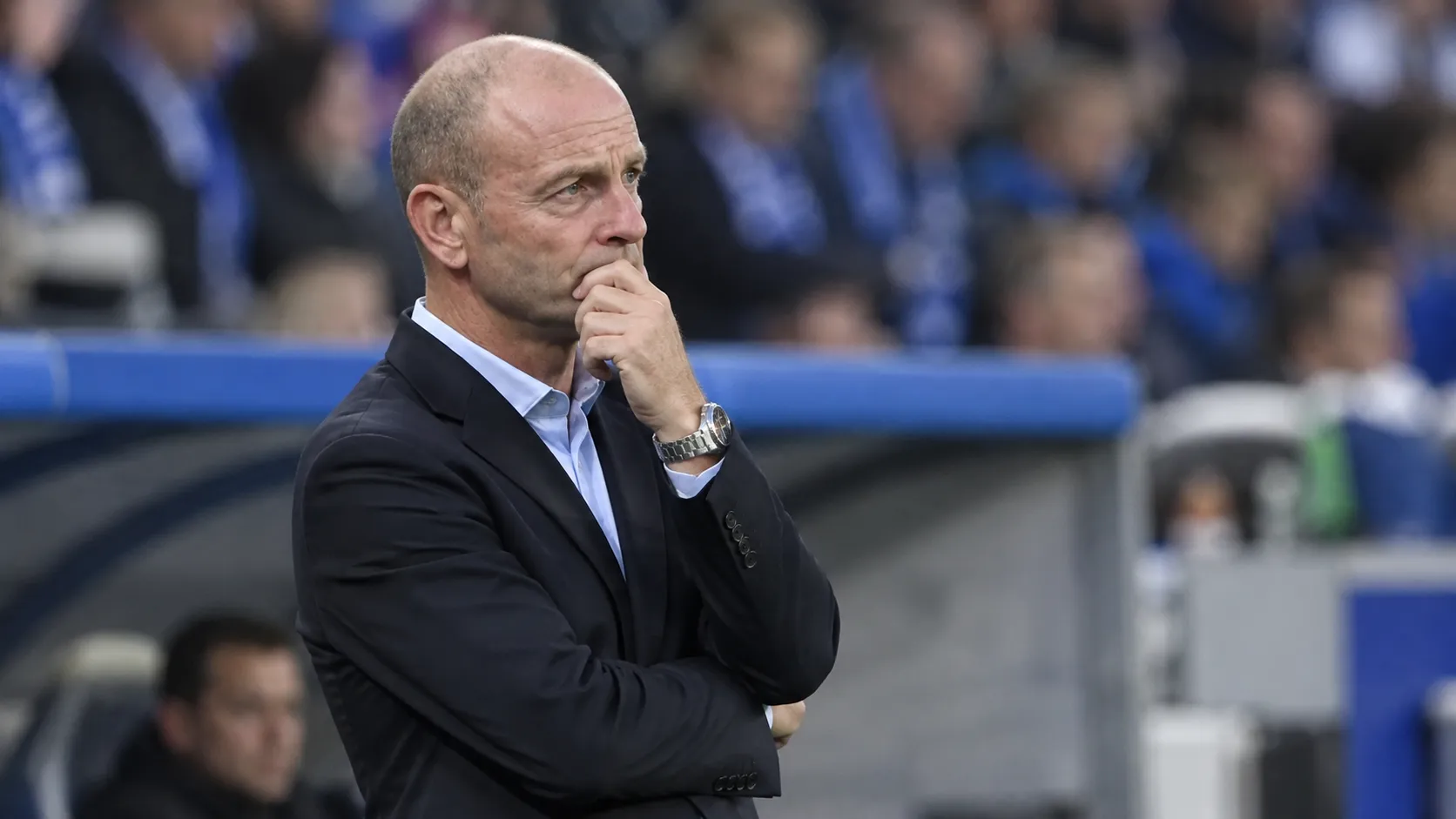 Een KRC Genk-coach in donker pak staat geconcentreerd langs de zijlijn.