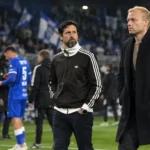 Medewerkers van Anderlecht staan langs de zijlijn terwijl spelers in blauwe shirts op de achtergrond lopen.